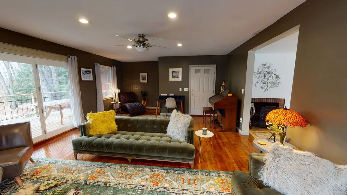 A cozy living room featuring plush green velvet sofas, a colorful area rug, a stylish lamp, and large windows with a view of the wooden deck. The decor suggests comfort and relaxation, while the view outside hints at the connection to nature and the balcony access described.