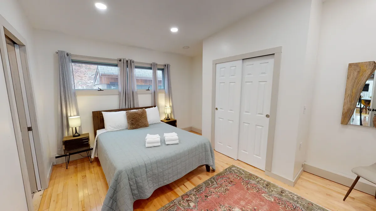 A bedroom with a light blue quilted bed, wooden nightstands, large windows letting in morning light, a soft rug, and double closet doors. The gentle color palette and soft textures evoke a sense of calm and comfort.