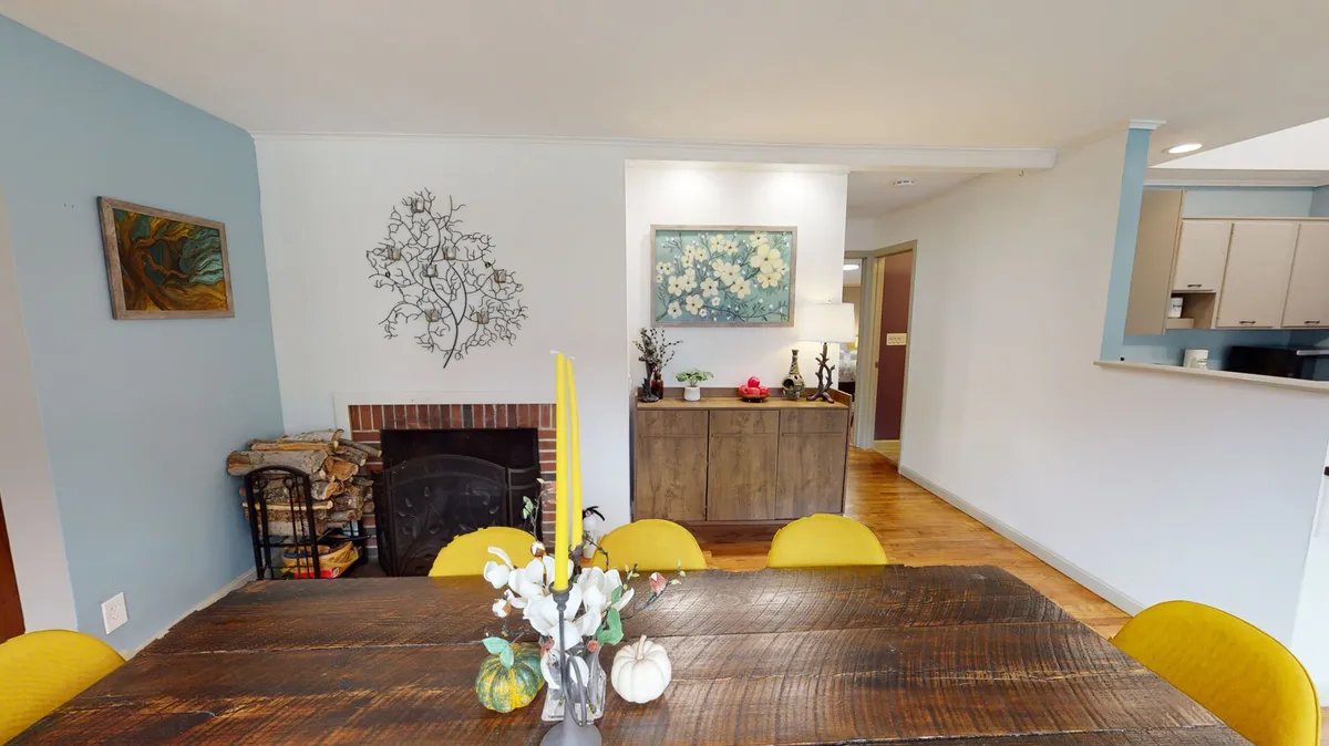 A warm, welcoming dining area with a stylish dining table, decorative wall art, a wood-burning fireplace, and bold yellow chairs. The scene suggests space for lively gatherings or intimate family dinners, with the fireplace adding a cozy, communal touch.