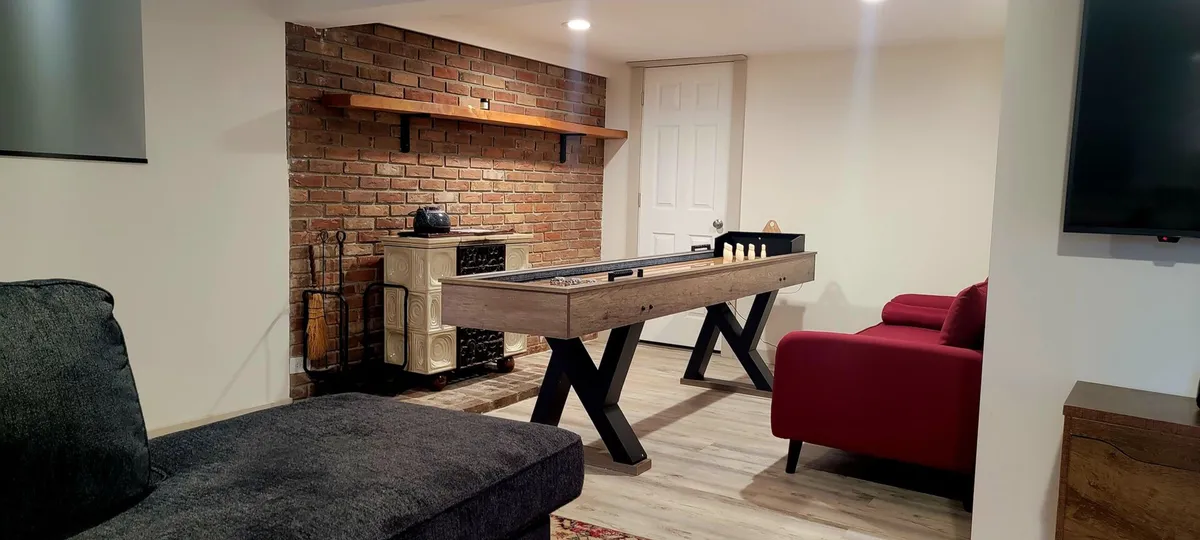 An interior game room with a shuffleboard table, playful red couch, exposed brick wall, and built-in shelves. The arrangement is ideal for group activities, fostering a lively, social atmosphere perfect for family tournaments and evening fun.