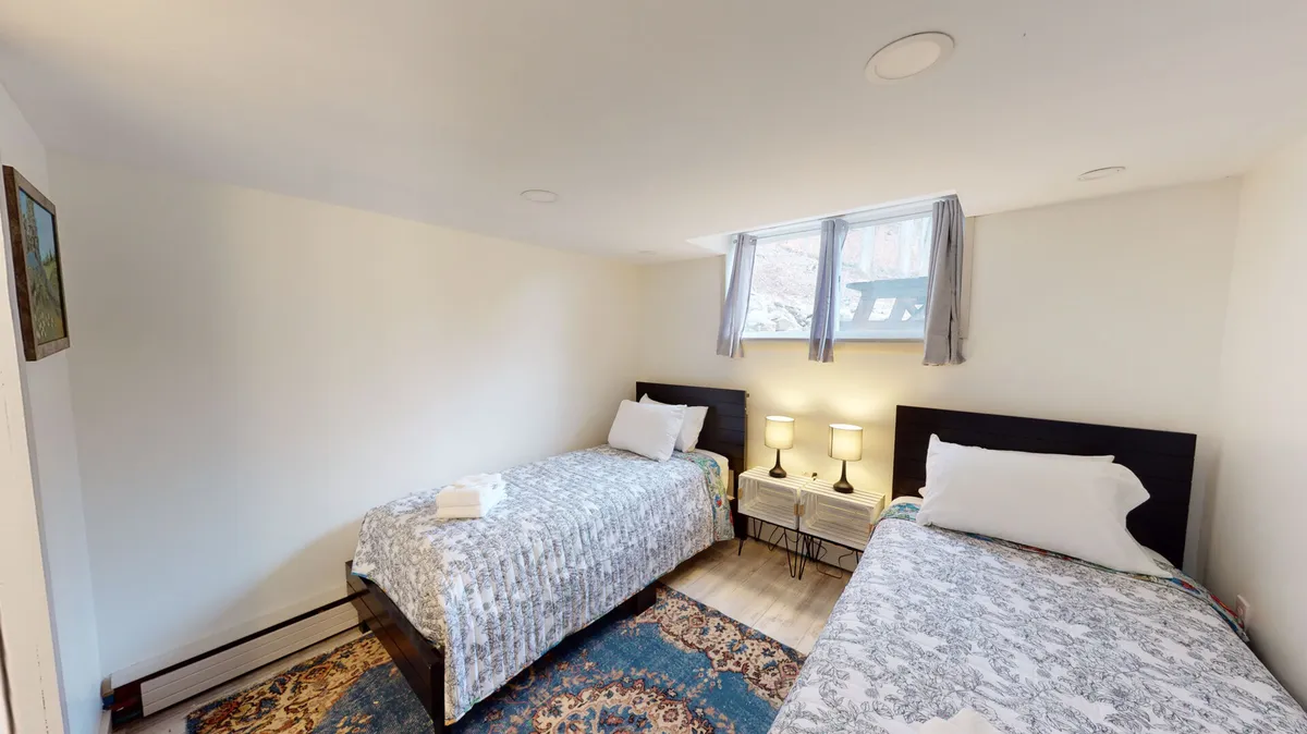 A cozy lower-level bedroom with two twin beds, a nightstand with lamps, and a curtained window. The setup is ideal for children or friends, with a relaxed mood and space for shared moments.