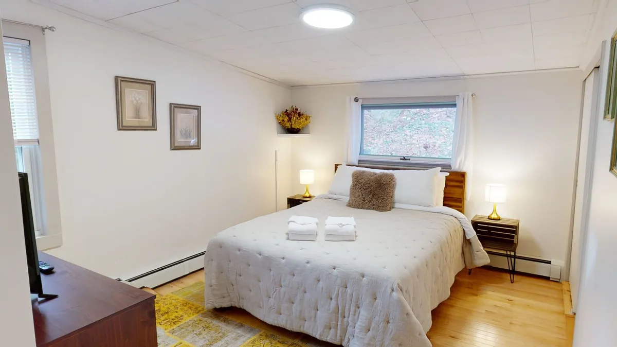A spacious bedroom featuring a plush bed, nightstands, a large window letting in natural light, decorative wall art, and a wall-mounted television. The décor is inviting and tranquil, with soft bedding and a cozy ambiance perfect for relaxation.
