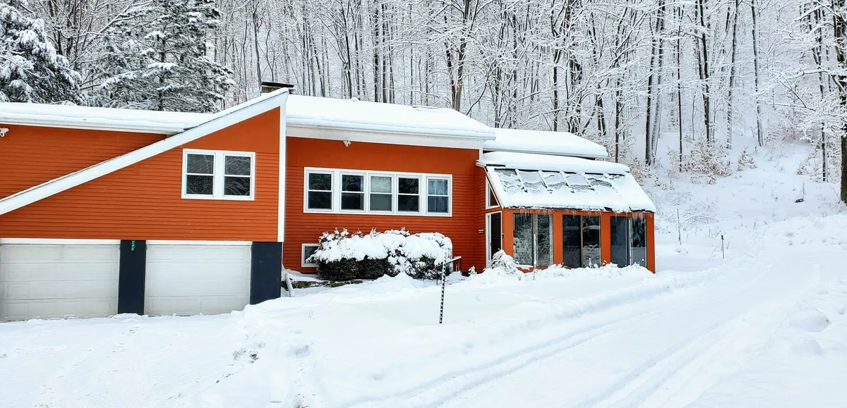 A winter scene featuring the house's bright orange exterior framed by a snowy landscape. Large windows and a sunroom with glass panels capture the light, while the outside area hints at winter sports and cozy gatherings inside. The mood is warm and inviting despite the cold surroundings, with subtle cues of outdoor play and relaxation.