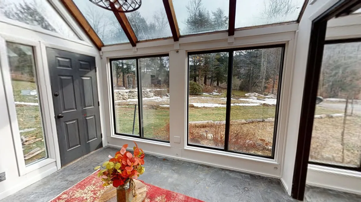 This image features a bright interior space with large windows that let in abundant natural light, offering views of the surrounding greenery. A colorful flower arrangement sits atop a textured area rug, contributing to a calm and serene mood. The black door and soft furnishings create a welcoming atmosphere, ideal for relaxation or enjoying quiet moments.