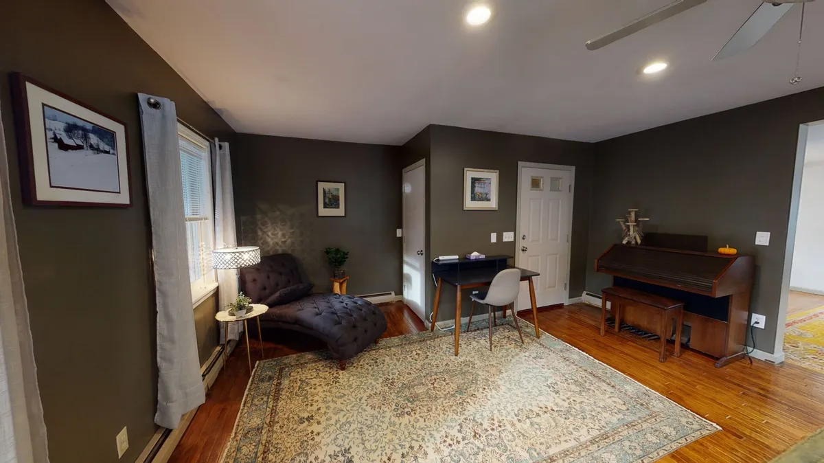 A cozy living space featuring a comfortable sofa, wooden dining table, upright piano, and a colorful rug. Natural light streams in through large windows, hinting at scenic views and a tranquil, inviting atmosphere ideal for both work and relaxation.